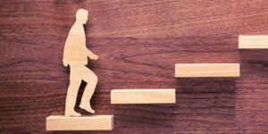 Wooden cutout of a person walking up a staircase made of wooden blocks, symbolising growth, progress, and personal or professional development.