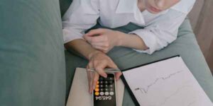 Person in a white shirt sitting on a couch, using a calculator while holding a pen.