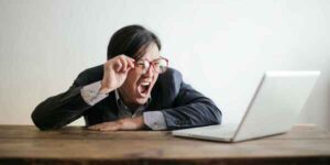 Frustrated man in a suit with red glasses, yelling at his laptop while sitting at a wooden table.