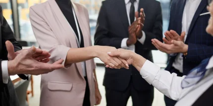 Two business people shaking hands while others clap